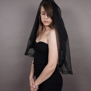 May include: A woman wearing a black strapless dress and a black veil. She is standing in front of a grey background.