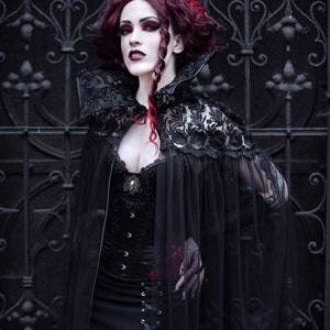May include: A woman in a black lace cape and corset stands in front of a black metal gate. Her hair is red and she has dark makeup on.