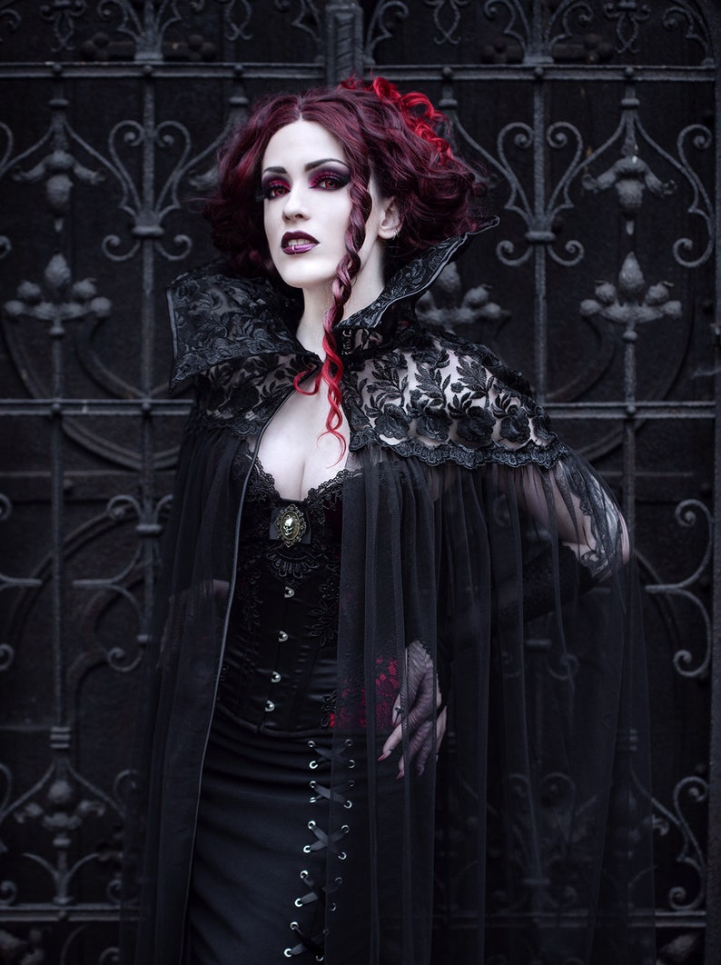May include: A woman in a black lace cape and corset stands in front of a black metal gate. Her hair is red and she has dark makeup on.