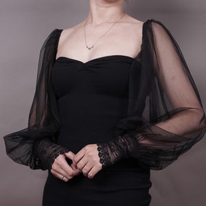 Könnte beinhalten: Ein schwarzes Kleid mit durchsichtigen, bauschigen Ärmeln und einer Spitzenborte an den Manschetten. Das Kleid hat einen herzförmigen Ausschnitt und ein figurbetontes Oberteil.