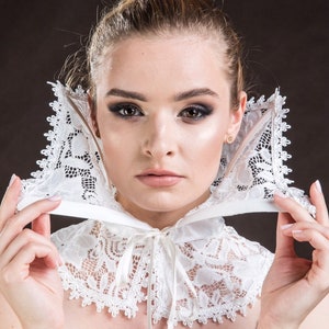 May include: A white lace collar with a satin ribbon tie. The collar is being worn by a person with blonde hair and dark eye makeup.
