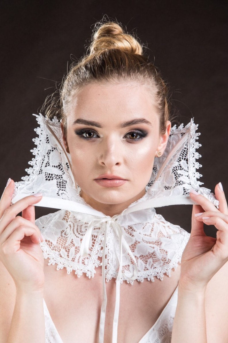 May include: A white lace collar with a satin ribbon tie. The collar is being worn by a person with blonde hair and dark eye makeup.