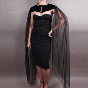 May include: A black strapless midi dress featuring a fitted bodice with a subtle twist detail at the center bust. The dress is paired with a black velvet bolero-style shrug that includes long, flowing, sheer tulle sleeves that drape to the floor.