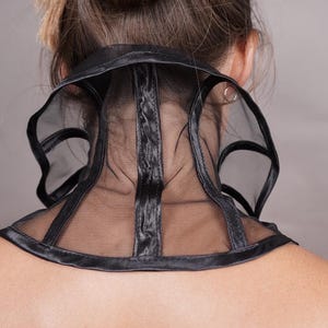 Pu&ograve; includere: Un accessorio da collo nero dal design strutturato, caratterizzato da pannelli in rete trasparente e bordi neri solidi. L'articolo avvolge il collo, mostrando una fascia centrale verticale e aperture laterali curve, creando un aspetto geometrico.