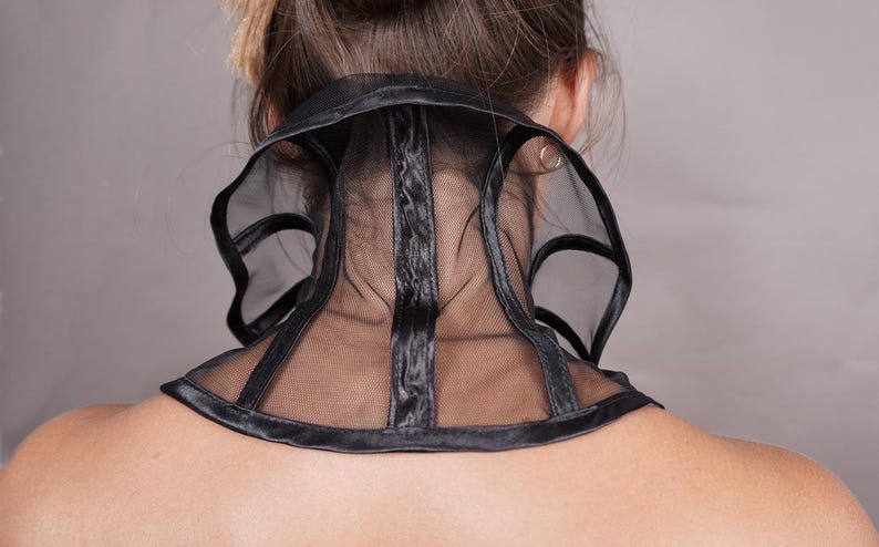 Pu&ograve; includere: Un accessorio da collo nero dal design strutturato, caratterizzato da pannelli in rete trasparente e bordi neri solidi. L'articolo avvolge il collo, mostrando una fascia centrale verticale e aperture laterali curve, creando un aspetto geometrico.