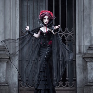 May include: A woman in a black gothic dress and cape with a red rose detail stands in front of a wrought iron gate. She has red hair and pale makeup.