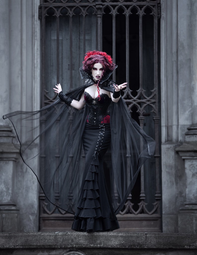 May include: A woman in a black gothic dress and cape with a red rose detail stands in front of a wrought iron gate. She has red hair and pale makeup.