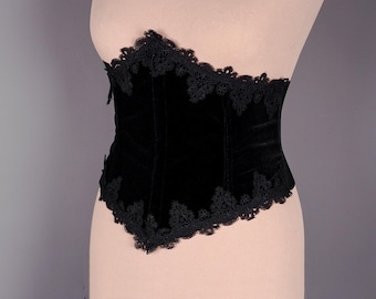 May include: A black velvet corset with black lace trim. The corset has a pointed shape and is designed to cinch the waist.