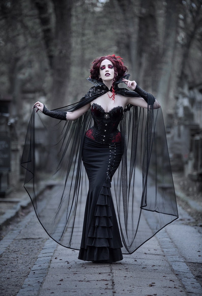 May include: A woman in a black gothic dress with a lace-trimmed cape and a red corset. She has red hair and is standing in a cemetery.