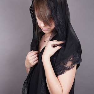 May include: A woman wearing a black lace shawl with a delicate gold necklace. The shawl is sheer and has a scalloped edge. The woman is looking down and her hair is partially covered by the shawl.