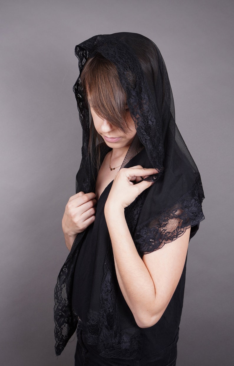 May include: A woman wearing a black lace shawl with a delicate gold necklace. The shawl is sheer and has a scalloped edge. The woman is looking down and her hair is partially covered by the shawl.