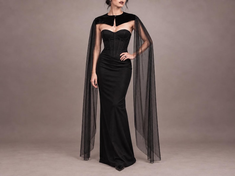 May include: A floor-length black strapless gown featuring a structured corset bodice with vertical seams. The dress is paired with a black velvet bolero-style shoulder wrap that includes long, flowing, sheer black tulle sleeves that extend to the floor.