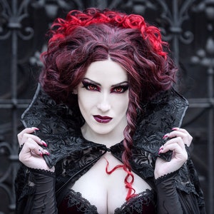 May include: A woman with long, dark red hair and red lipstick wears a black lace cape with a high collar. She has dark eye makeup and red nail polish.