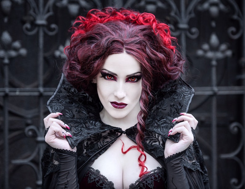 May include: A woman with long, dark red hair and red lipstick wears a black lace cape with a high collar. She has dark eye makeup and red nail polish.