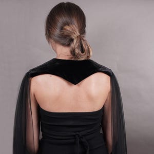 May include: A black velvet shoulder wrap featuring long, sheer black tulle sleeves that drape down the arms. The garment is worn over a black strapless top with a tie-back closure, creating a layered look with contrasting fabric textures.