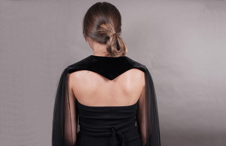 May include: A black velvet shoulder wrap featuring long, sheer black tulle sleeves that drape down the arms. The garment is worn over a black strapless top with a tie-back closure, creating a layered look with contrasting fabric textures.