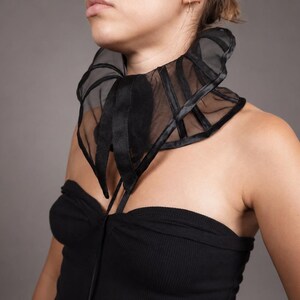 Pu&ograve; includere: Un colletto nero in organza trasparente con stecche verticali strutturate e chiusura con nastro di raso sul davanti. L'accessorio si indossa intorno al collo e presenta un design a collo alto con volant, cuciture e bordi in evidenza.