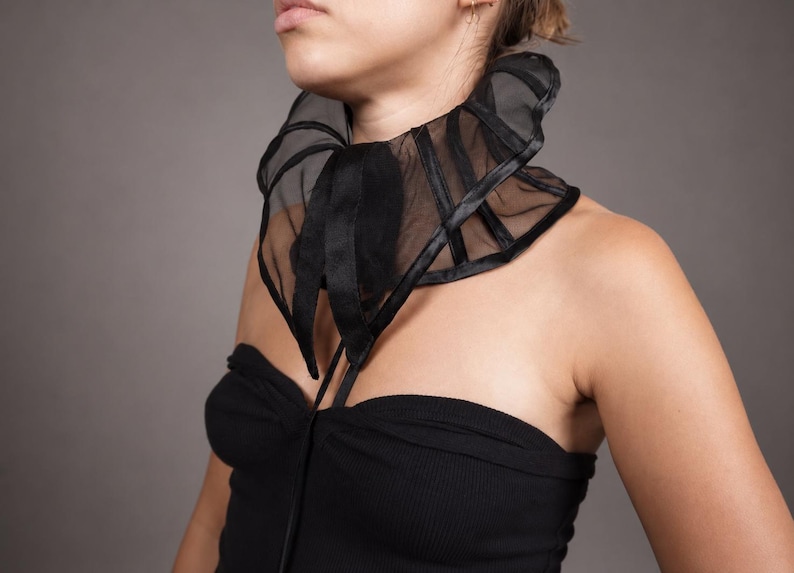 Pu&ograve; includere: Un colletto nero in organza trasparente con stecche verticali strutturate e chiusura con nastro di raso sul davanti. L'accessorio si indossa intorno al collo e presenta un design a collo alto con volant, cuciture e bordi in evidenza.