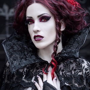 May include: A woman with long, dark red hair styled in curls and braids wears a black lace cape with a high collar. She has dramatic makeup with red eyeshadow and red contact lenses.