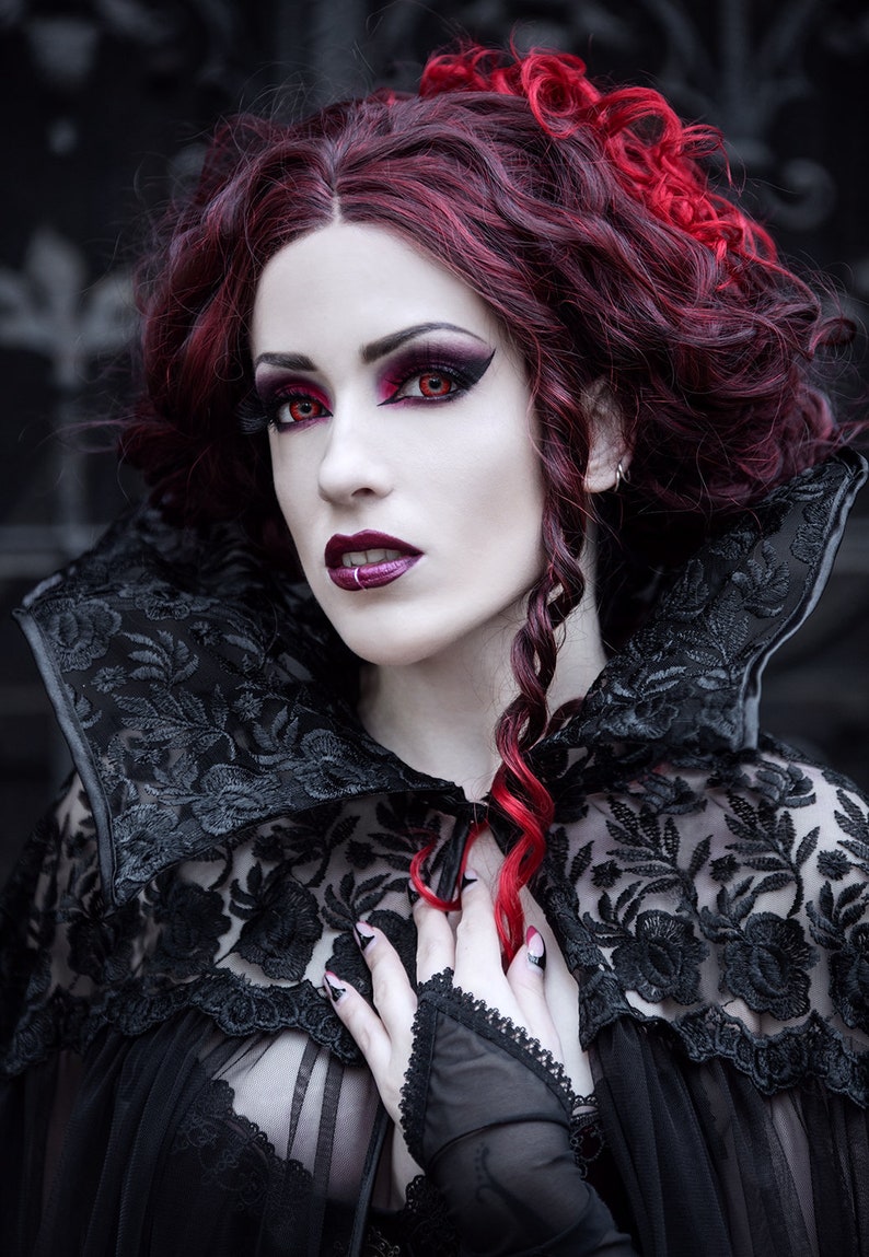 May include: A woman with long, dark red hair styled in curls and braids wears a black lace cape with a high collar. She has dramatic makeup with red eyeshadow and red contact lenses.