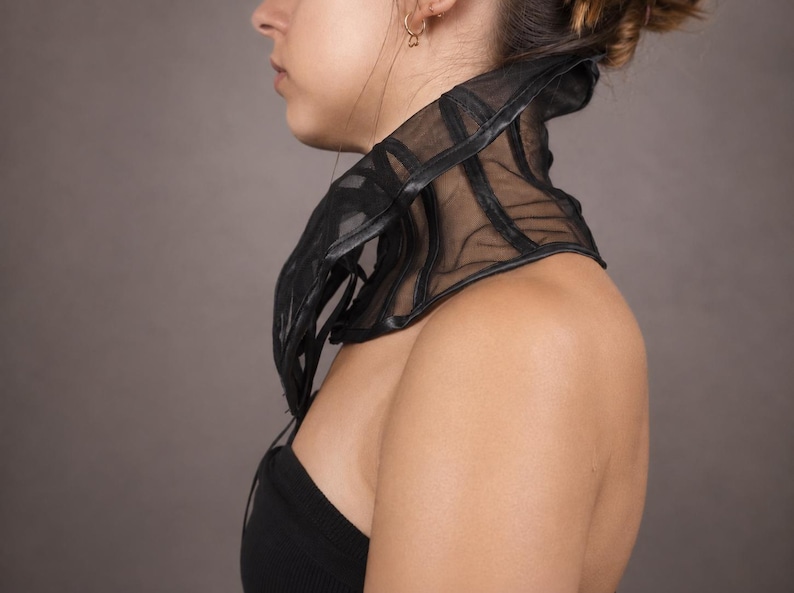 Pu&ograve; includere: Un accessorio per il collo in rete nera trasparente con stecche strutturate e bordi scuri. Il pezzo avvolge il collo e la parte superiore del torace, creando una silhouette a collo alto con cuciture verticali visibili e tessuto semitrasparente.