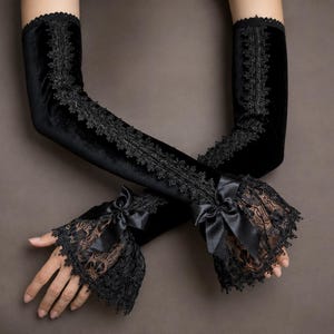 May include: A pair of black velvet opera-length gloves featuring a vertical strip of intricate black lace running down the centre. The cuffs are adorned with a ruffled brown lace trim, a black satin ribbon bow, and a scalloped black lace edge.