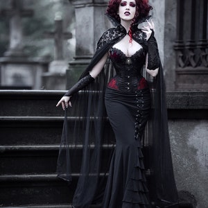 May include: A woman in a black lace cape and a black corset with red accents stands on a set of stone steps. She has red hair and is holding a small object in her hand.