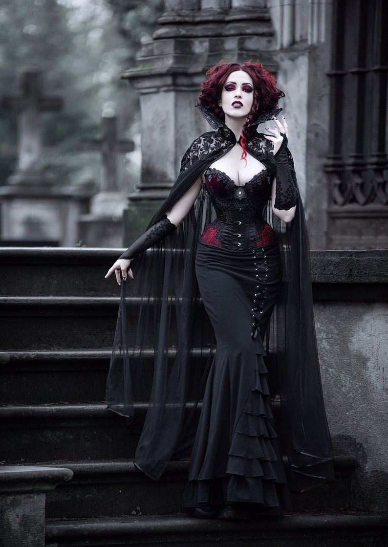 May include: A woman in a black lace cape and a black corset with red accents stands on a set of stone steps. She has red hair and is holding a small object in her hand.