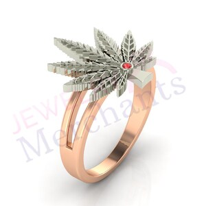 May include: A gold ring with a three-dimensional marijuana leaf design. The leaf is made of white gold and has a red gemstone in the center. The ring band is made of rose gold.