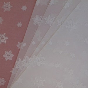 May include: A stack of white tissue paper with a snowflake pattern. The tissue paper is folded in half and the top layer is slightly offset from the bottom layer. The tissue paper is a light pink color.