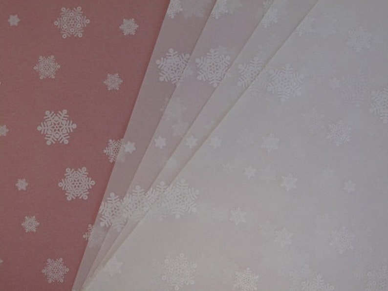May include: A stack of white tissue paper with a snowflake pattern. The tissue paper is folded in half and the top layer is slightly offset from the bottom layer. The tissue paper is a light pink color.