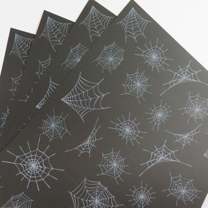 May include: Four sheets of black paper featuring a repeating pattern of light blue spiderwebs. The spiderwebs vary in size and design, with some appearing as complete webs and others as partial or abstract shapes. The paper is fanned out, showing the pattern.