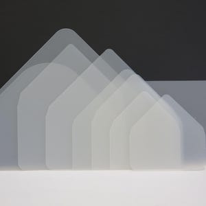 Op de afbeelding: Een verzameling doorschijnende, huisvormige plastic sjablonen gerangschikt in een gelaagd patroon. De sjablonen zijn in verschillende maten, met afgeronde toppen en rechte zijkanten, waardoor een gradiënteffect ontstaat.