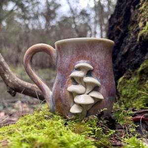 May include: A ceramic mug with a glossy, multi-coloured glaze in shades of purple, brown, and blue. The exterior features a three-dimensional, sculpted cluster of off-white mushrooms attached to the side, complemented by a curved handle.