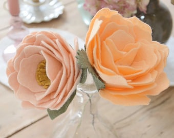 May include: Two peach-colored felt flowers in a clear glass vase on a wooden table. The flowers have a layered petal design.