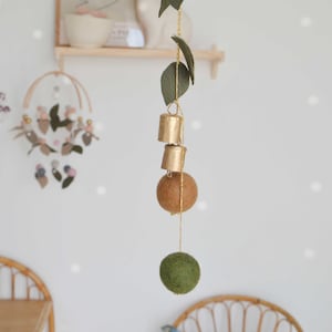 Könnte beinhalten: Eine goldene und grüne Filzgirlande mit drei goldenen Glöckchen und zwei Filzkugeln, die an einer goldenen Schnur hängen. Die Girlande hängt von einer Decke in einem Raum mit weißen Wänden und einem Holzregal.