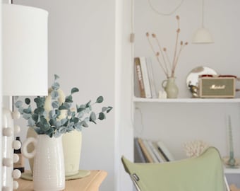 May include: A white ceramic pitcher with a handle sits on a wooden table with a white lamp and a vase of eucalyptus leaves. The pitcher is on a wooden table with a white lamp and a vase of eucalyptus leaves.