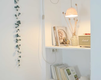 May include: A white shelf with a white ceramic lampshade hanging from the ceiling. The shelf is decorated with books, a vase with dried flowers, and a small bird figurine. The wall is white with a green garland hanging on the left side.