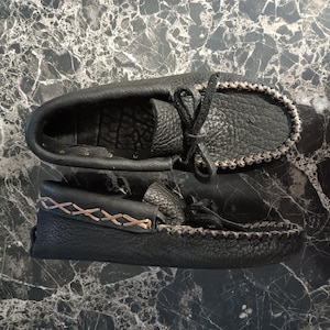 May include: A pair of black leather moccasins featuring a textured grain finish, a decorative whipstitch detail along the collar in a light tan thread, and a matching leather lace tie centered on the top of the vamp for a secure fit.