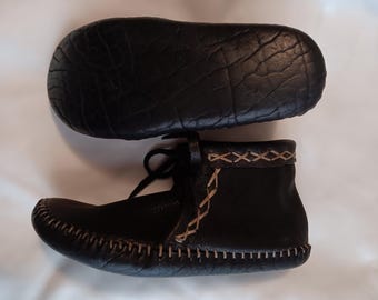 May include: Black leather moccasins with a brown stitching detail around the sole and top of the shoe. The moccasins have a lace-up closure.