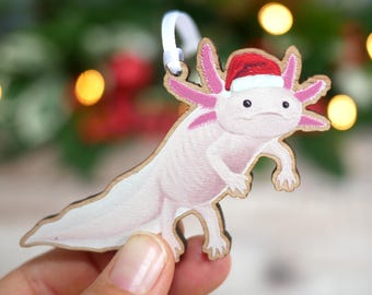 Könnte beinhalten: Ein flacher Axolotl-Anhänger aus Holz mit einem blassrosa Körper, dunkelrosa Kiemen und einer kleinen rot-weißen Weihnachtsmütze. Der Anhänger ist mit einer weißen Bandschlaufe an der Oberseite zum Aufhängen versehen.