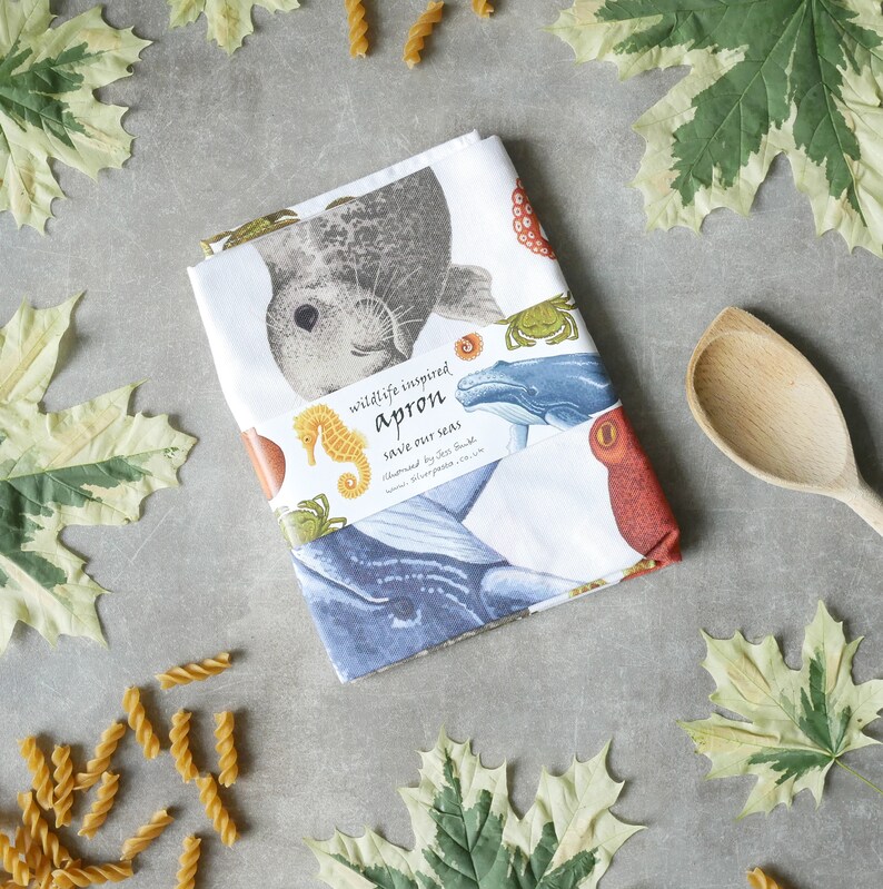 Pu&ograve; includere: Grembiule bianco con illustrazioni di vita marina, tra cui una foca, un cavalluccio marino e una balena. Il grembiule ha il testo "wildlife inspired apron" e "save our seas". Un cucchiaio di legno e pasta sullo sfondo.