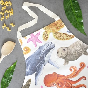 Pu&ograve; includere: Grembiule bianco con illustrazioni colorate di vita marina, tra cui una balena, un polpo, una tartaruga marina, una stella marina e una foca. Il grembiule ha un cinturino e lacci color crema. Misure in centimetri.