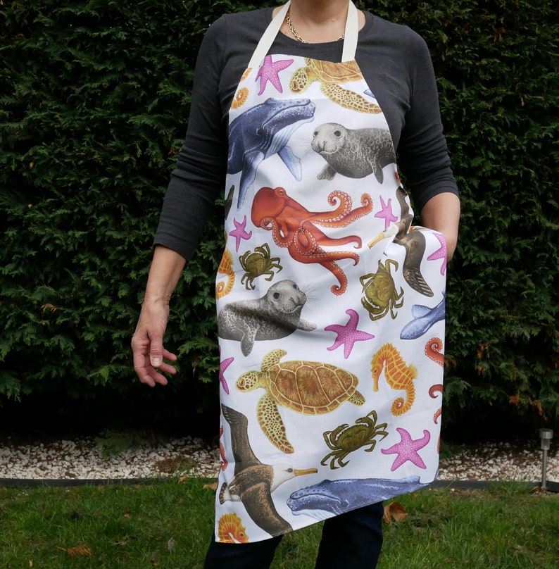 Pu&ograve; includere: Grembiule bianco con un vivace motivo di vita marina. Il design include illustrazioni di balene, foche, polpi, tartarughe marine, stelle marine, granchi e cavallucci marini. Ha una tasca e spalline color crema.