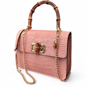 Puede incluir: Bolso de mano rosa con textura de cocodrilo, asa de bambú y correa de cadena dorada. El bolso tiene forma rectangular con cierre de solapa y herrajes dorados. Medidas no visibles.