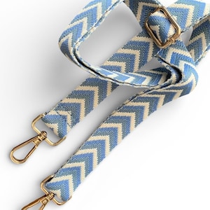 May include: A woven bag strap featuring a blue and white chevron pattern. The strap includes gold-toned metal hardware, including two lobster claw clasps at the ends and an adjustable sliding buckle to modify the length of the strap as needed.