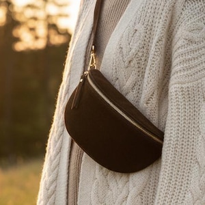 May include: A dark brown fanny pack with a gold zipper and strap is worn across the body. The bag is made of a soft material. The person is wearing a cream-colored cable knit cardigan and a beige sweater. The background is a field with trees.