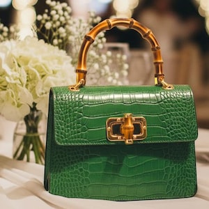 May include: A vibrant green handbag with a textured crocodile-embossed finish, featuring a distinctive arched bamboo handle and a gold-toned metal clasp closure. The structured bag is positioned on a white fabric surface.