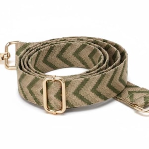 Puede incluir: Una correa de bolso ajustable enrollada con un patrón de chevrón en color tostado y verde oliva. La correa incluye herrajes de metal en tono dorado pulido, con una hebilla deslizante rectangular para ajustar la longitud y dos mosquetones giratorios.