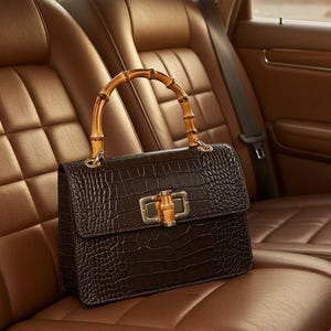Puede incluir: Un bolso marrón oscuro con estampado de cocodrilo descansa sobre un asiento de coche de cuero marrón. El bolso presenta un asa de bambú y un cierre dorado. El interior del coche tiene asientos de cuero marrón.